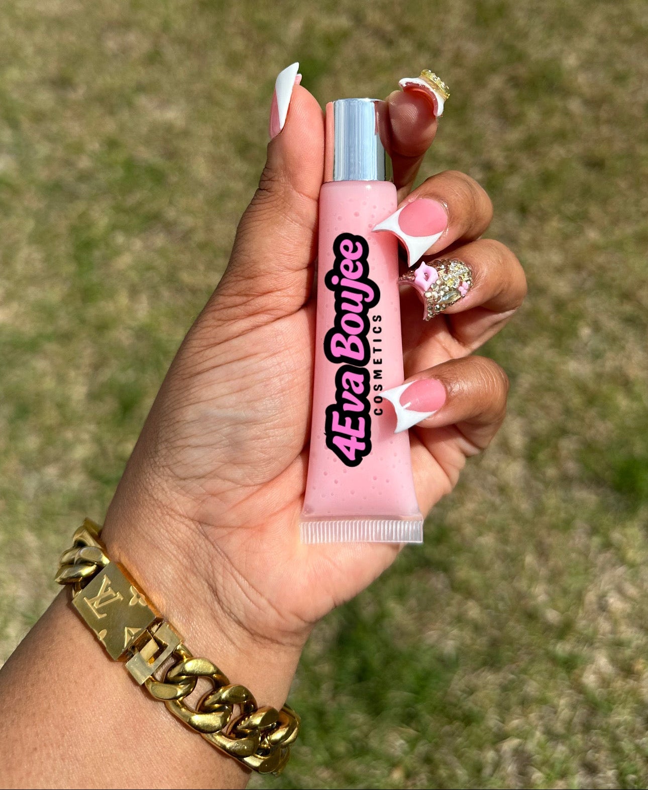 Hand holding a pink lip gloss with '4Ever Boujee' text against a blurred grass background