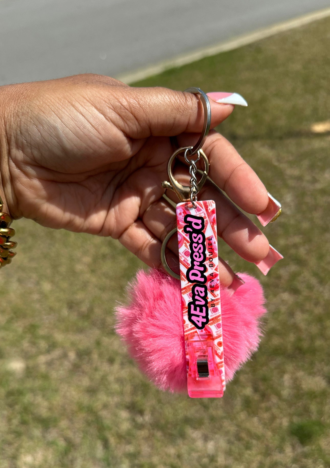 Hand holding a pink keychain with fluffy attachment against a blurred grassy background