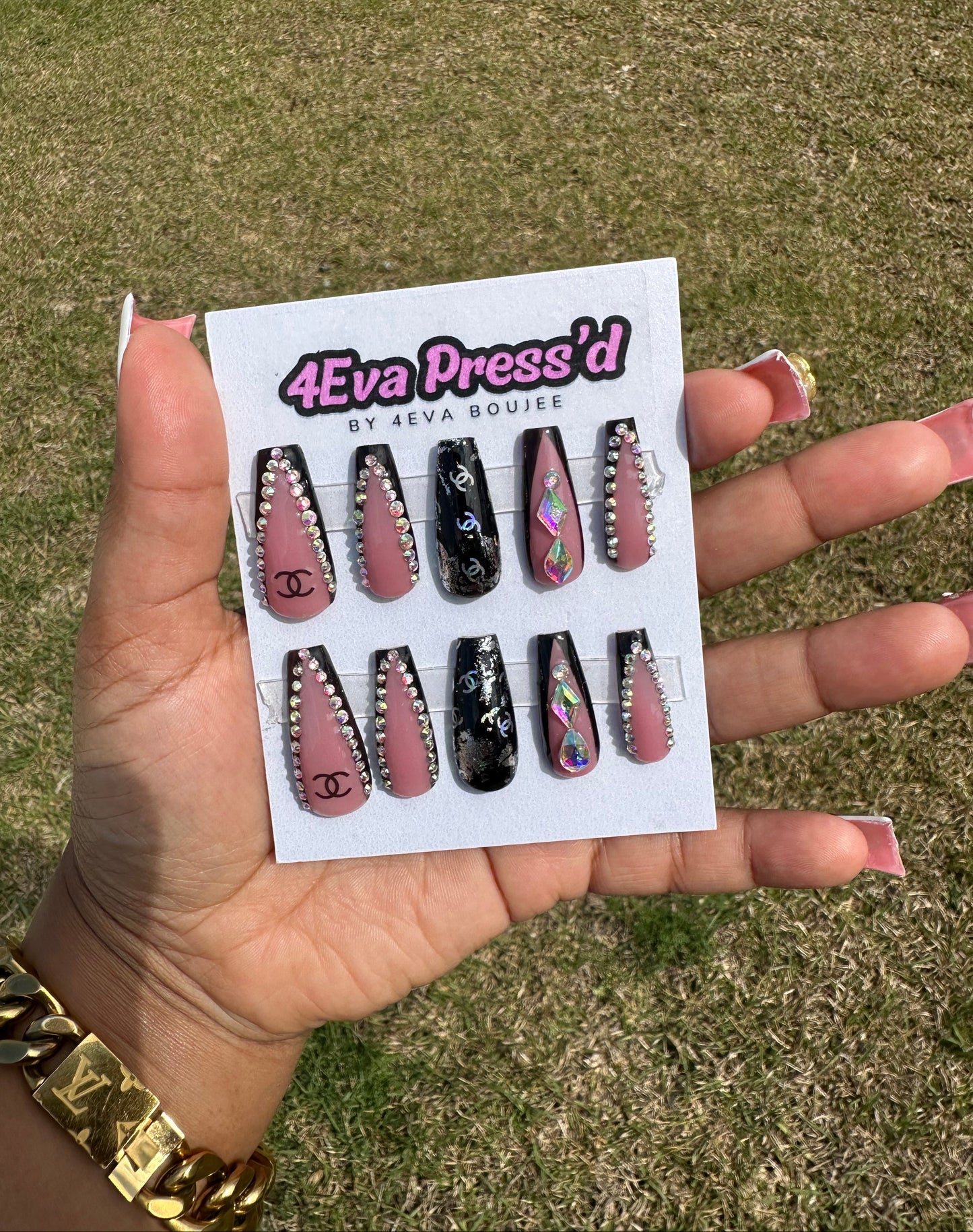 Hand holding a card with false nails labeled '4Eva Press'd' against a grassy background. 