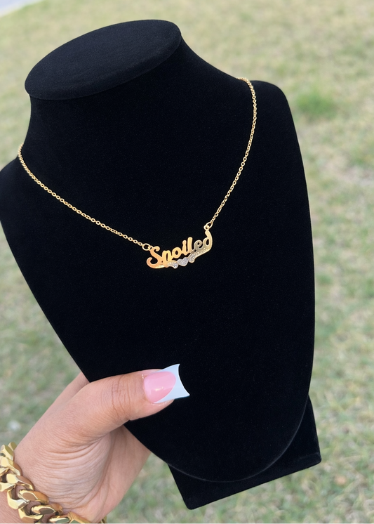Gold necklace with 'Spoiled' on a black stand held by a hand against a grassy background